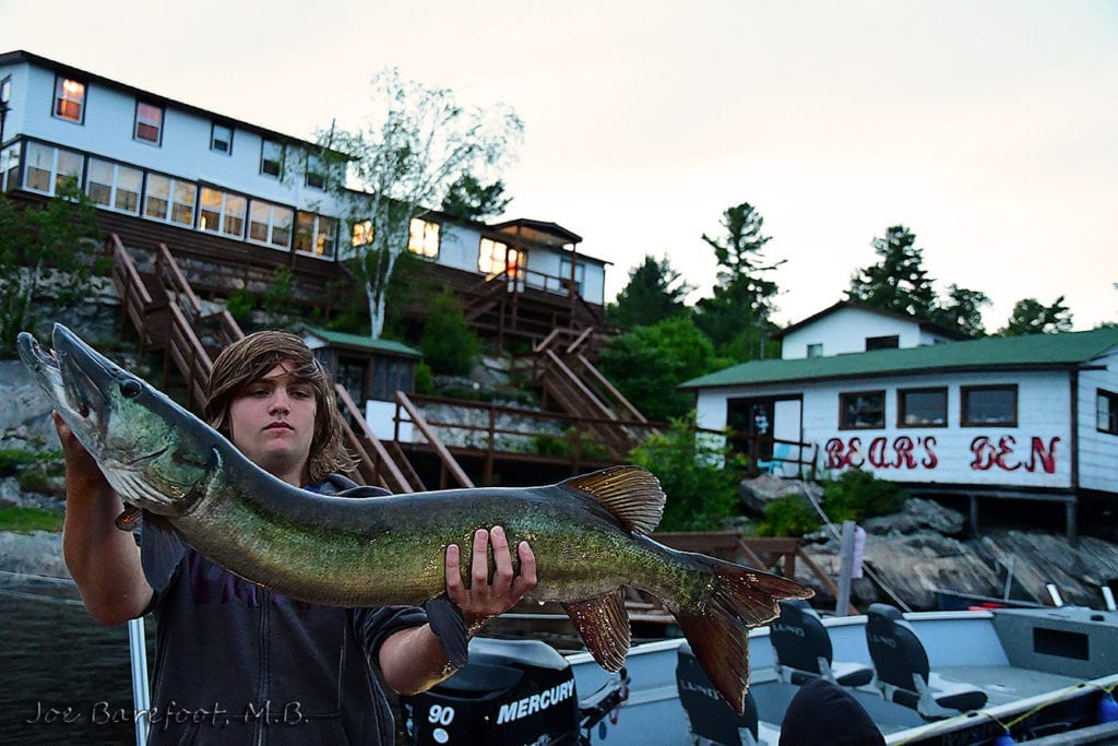 Mathias holding a 46 inch Musky with Bear's Den Lodge in background, Image captured by Joe Barefoot on Nikon D500