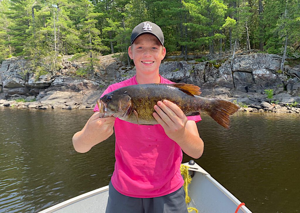 Image features rocky structure with a boy holding a French River Smallmouth Bass.